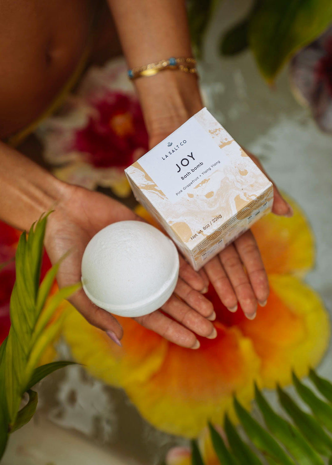 A woman holding a Joy Bath Bomb in a floral bath